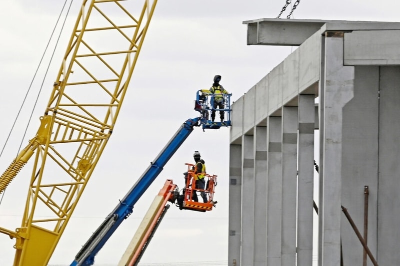 A gray concrete building framework under construction against an overcast sky. On the left, a large yellow crane is visible alongside two workers in high-visibility vests operating blue and orange boom lifts.