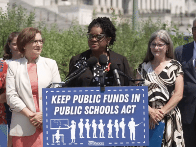 U.S. Rep. Gwen Moore speaks at an outdoor podium while surrounded by four colleagues. She stands behind a prominent blue sign with white text and a classroom graphic that reads, "KEEP PUBLIC FUNDS IN PUBLIC SCHOOLS ACT."