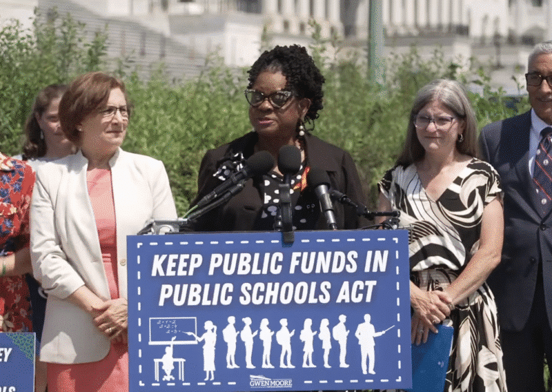 U.S. Rep. Gwen Moore speaks at an outdoor podium while surrounded by four colleagues. She stands behind a prominent blue sign with white text and a classroom graphic that reads, "KEEP PUBLIC FUNDS IN PUBLIC SCHOOLS ACT."