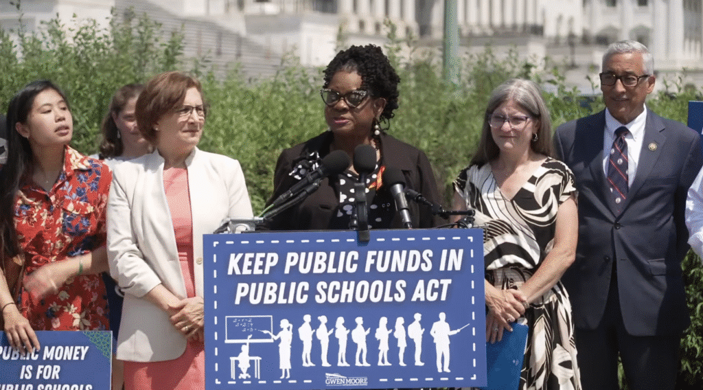 U.S. Rep. Gwen Moore speaks at an outdoor podium while surrounded by four colleagues. She stands behind a prominent blue sign with white text and a classroom graphic that reads, "KEEP PUBLIC FUNDS IN PUBLIC SCHOOLS ACT."