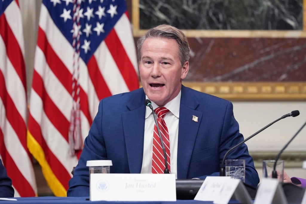 U.S. Senator Jon Husted sits at a table speaking into a microphone while wearing a blue suit, white shirt, and a red-and-white striped tie. Two American flags stand in the background.