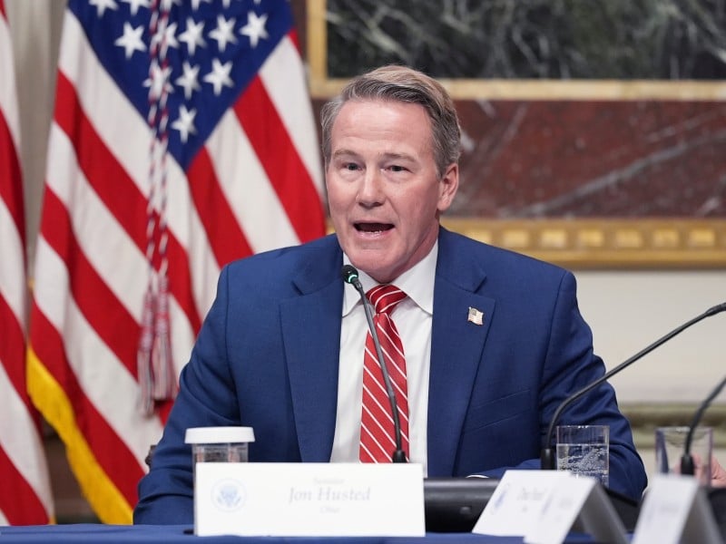 U.S. Senator Jon Husted sits at a table speaking into a microphone while wearing a blue suit, white shirt, and a red-and-white striped tie. Two American flags stand in the background.