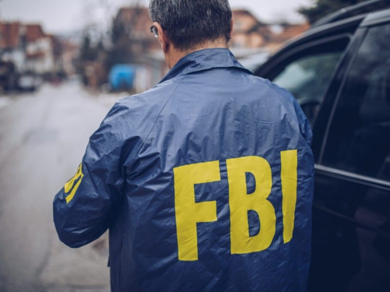 A person is viewed from behind standing next to a black car on a street. They are wearing a blue jacket with the text "FBI" printed in large yellow letters across the back