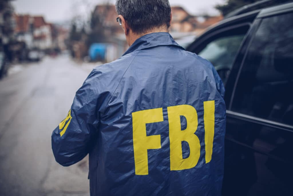 A person is viewed from behind standing next to a black car on a street. They are wearing a blue jacket with the text "FBI" printed in large yellow letters across the back