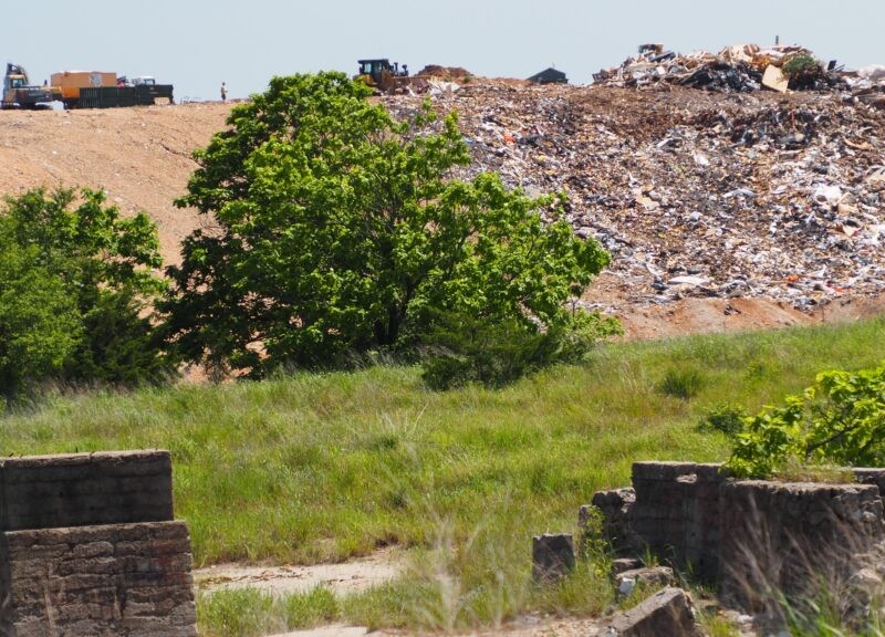 A massive mound of multicolored trash sits next to a tan dirt hill under a pale blue sky. In the foreground, vibrant green grass and trees grow around old concrete ruins. Heavy yellow construction vehicles are parked along the distant ridge of the landfill.