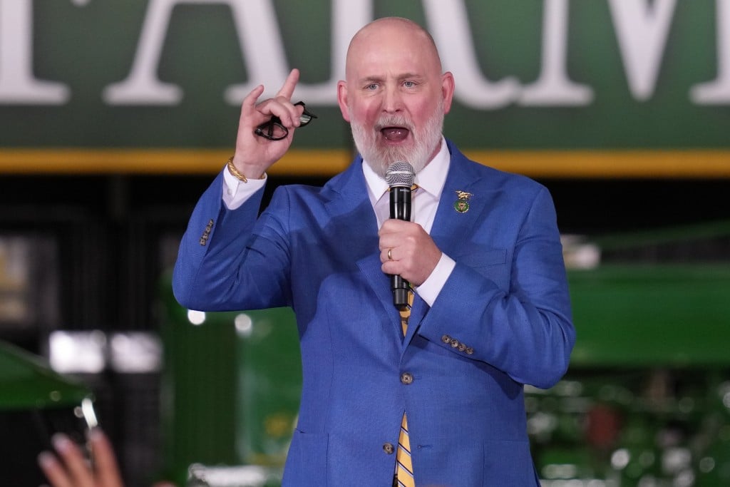U.S. Rep. Derrick Van Orden, a bald man with a white beard speaks into a microphone while wearing a bright blue suit jacket and a yellow-and-blue striped tie.