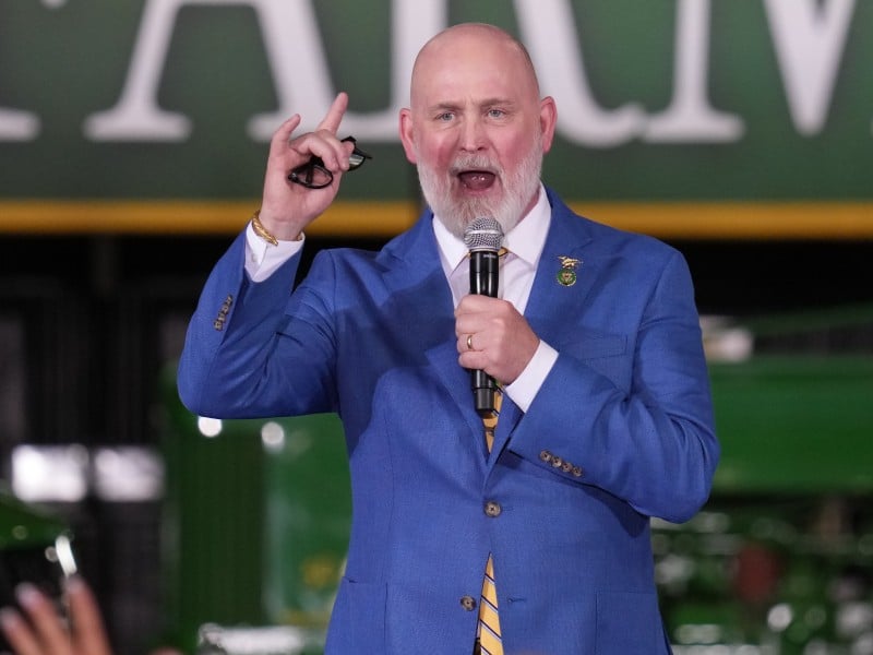 U.S. Rep. Derrick Van Orden, a bald man with a white beard speaks into a microphone while wearing a bright blue suit jacket and a yellow-and-blue striped tie.