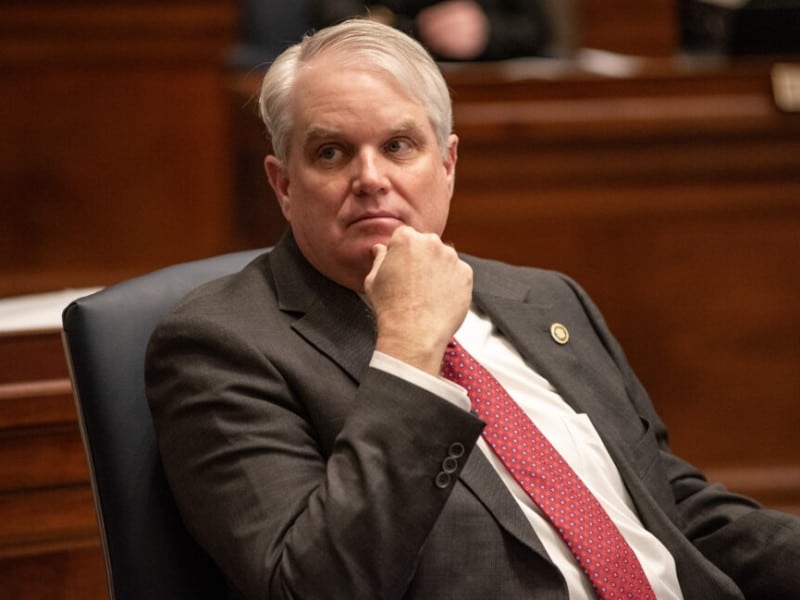 Missouri State Senator Jason Bean sits in a dark leather chair with his hand resting thoughtfully on his chin. He wears a dark grey suit jacket, a white shirt, and a patterned red tie.