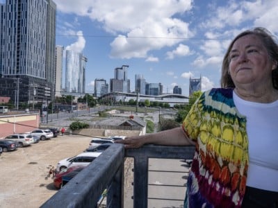 Tennessee resident Cassandra Boyle, a woman wearing a vibrant, multicolored tie-dye cardigan over a white shirt stands on an elevated balcony. She rests her hand on a metal railing against a backdrop of a city skyline filled with modern skyscrapers.