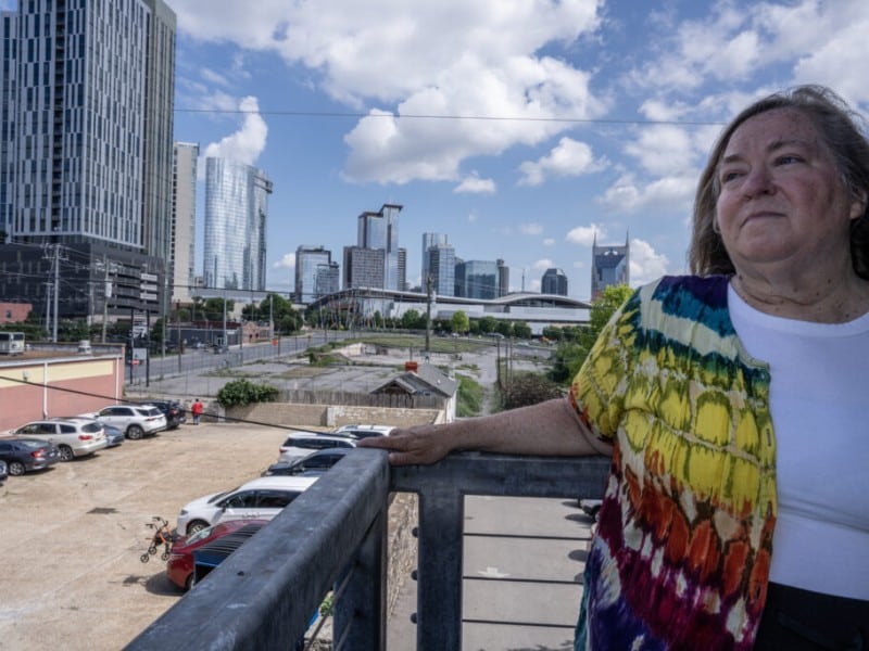 Tennessee resident Cassandra Boyle, a woman wearing a vibrant, multicolored tie-dye cardigan over a white shirt stands on an elevated balcony. She rests her hand on a metal railing against a backdrop of a city skyline filled with modern skyscrapers.