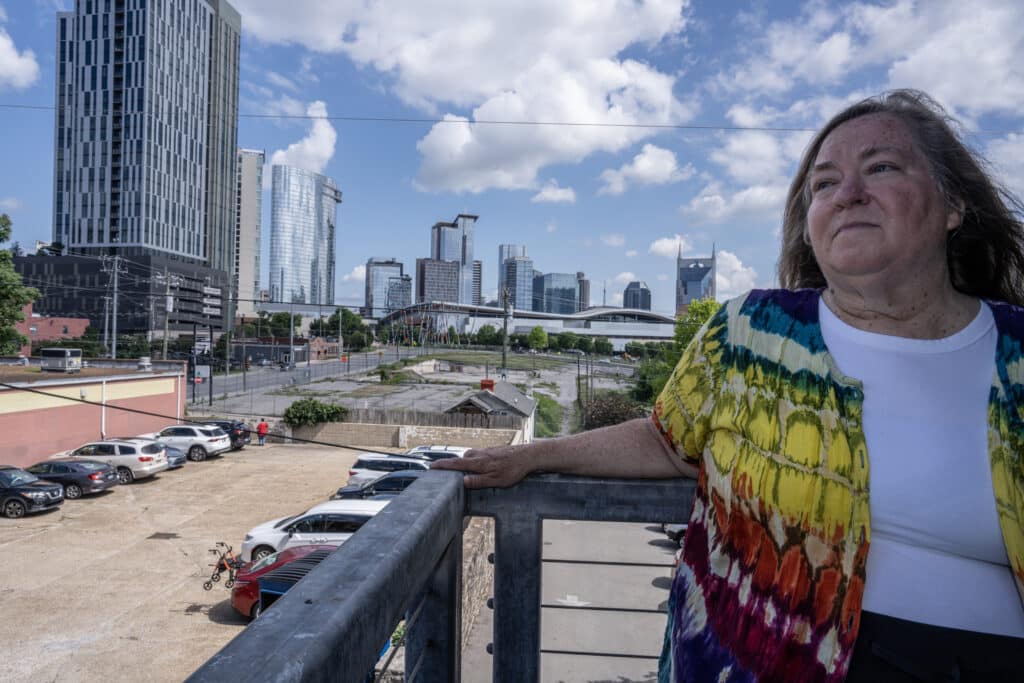 Tennessee resident Cassandra Doyle, a woman wearing a vibrant, multicolored tie-dye cardigan over a white shirt stands on an elevated balcony. She rests her hand on a metal railing against a backdrop of a city skyline filled with modern skyscrapers.