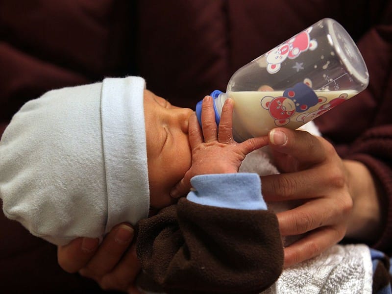 A close-up shows a newborn baby wearing a light blue beanie while being bottle-fed by an adult against a dark maroon background. The clear plastic bottle contains milk and features colorful red and blue cartoon bear graphics alongside printed measurement markings.