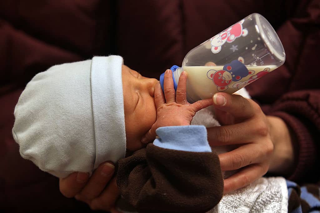 A close-up shows a newborn baby wearing a light blue beanie while being bottle-fed by an adult against a dark maroon background. The clear plastic bottle contains milk and features colorful red and blue cartoon bear graphics alongside printed measurement markings.