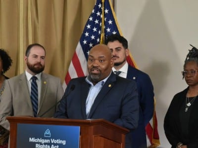 Democratic Michigan state Rep. Jason Hoskins is surrounded by colleagues and speaks at a podium with a sign that signs, "Michigan Voting Rights Act."