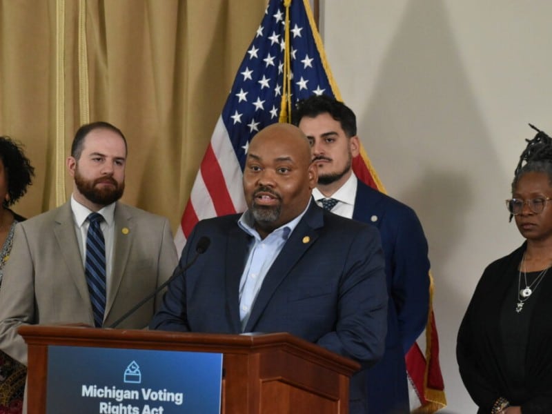 Democratic Michigan state Rep. Jason Hoskins is surrounded by colleagues and speaks at a podium with a sign that signs, "Michigan Voting Rights Act."