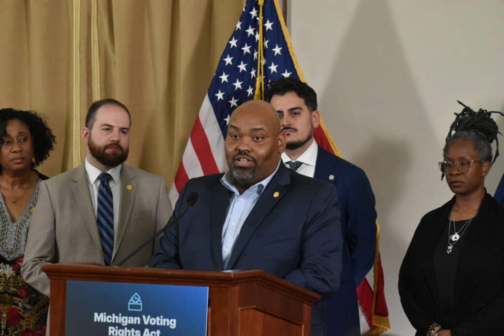 Democratic Michigan state Rep. Jason Hoskins is surrounded by colleagues and speaks at a podium with a sign that signs, "Michigan Voting Rights Act."
