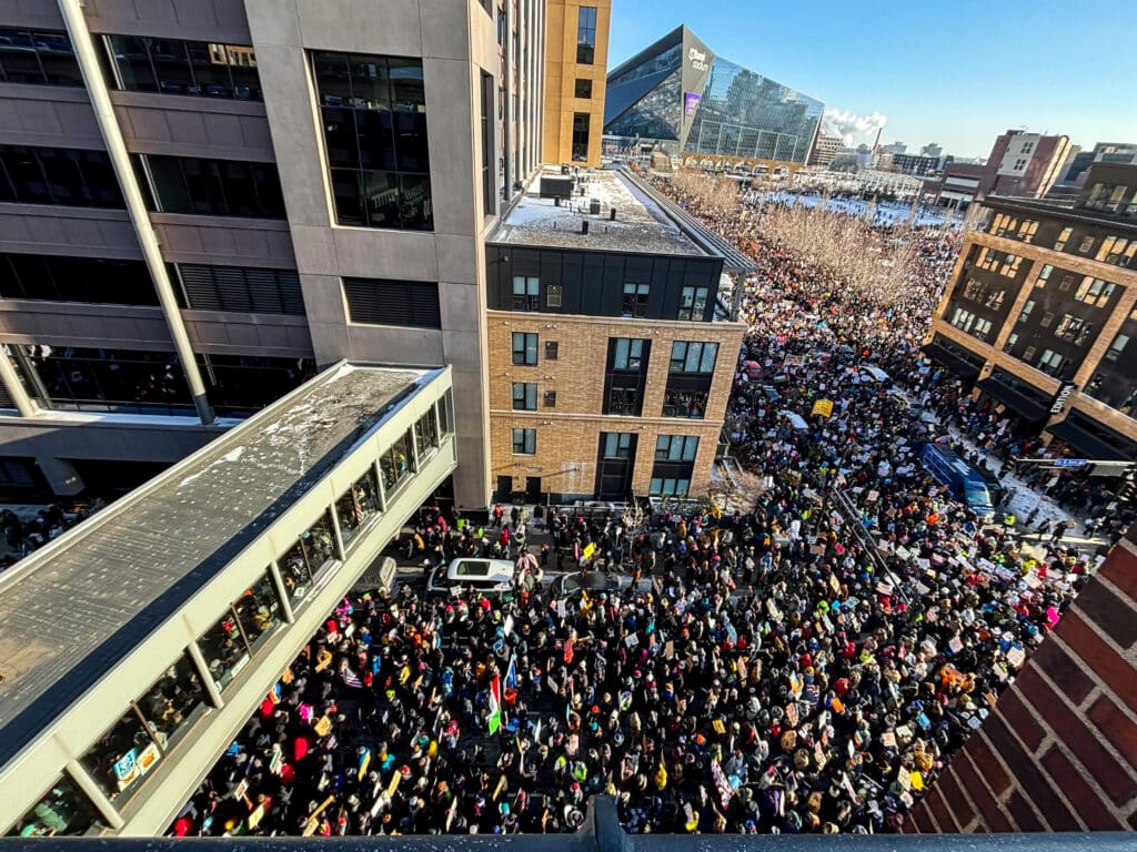 A high-angle view shows a massive crowd protesting on a city street in Minneapolis, Minnesota. People carry colorful signs and flags amidst patches of snow. In the distance, the U.S. Bank Stadium is visible under a clear blue sky.