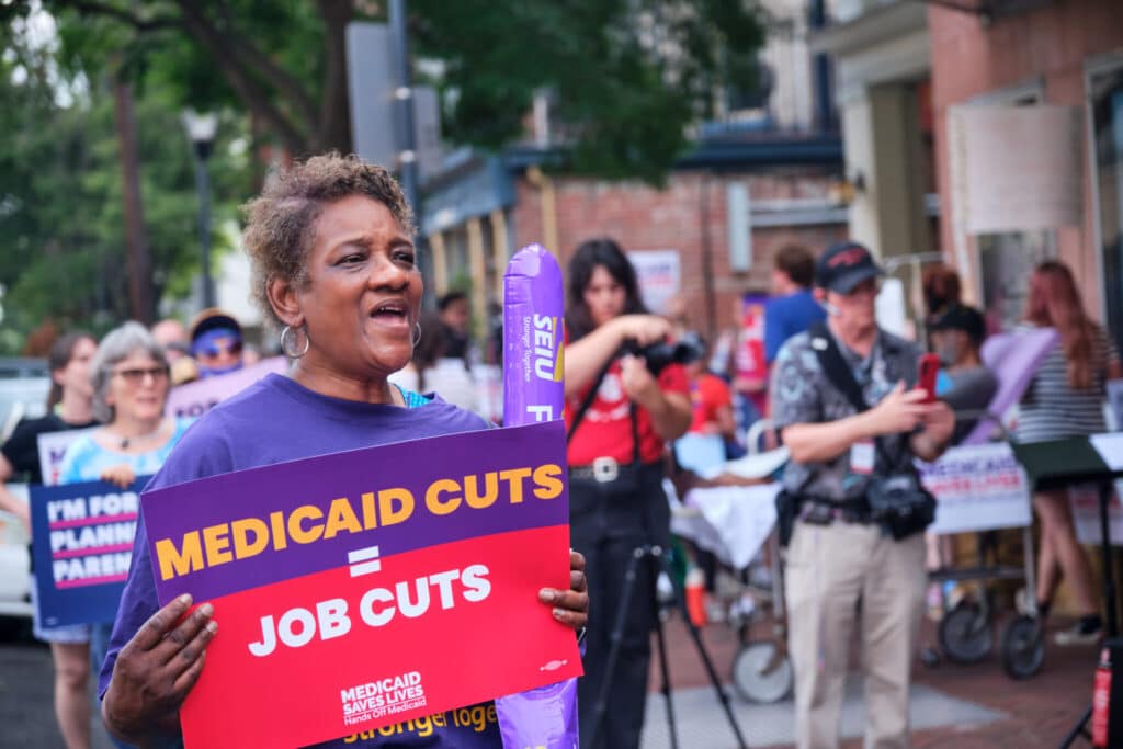 A demonstrator holds a sign reading "MEDICAID CUTS = JOB CUTS" at a rally.
