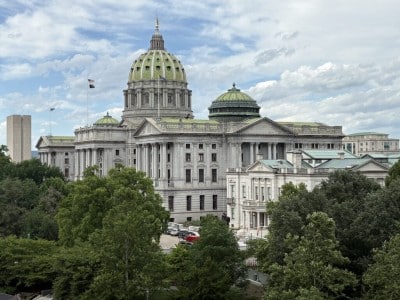 The Pennsylvania State Capitol building with its prominent green dome rises behind a line of dense green trees.