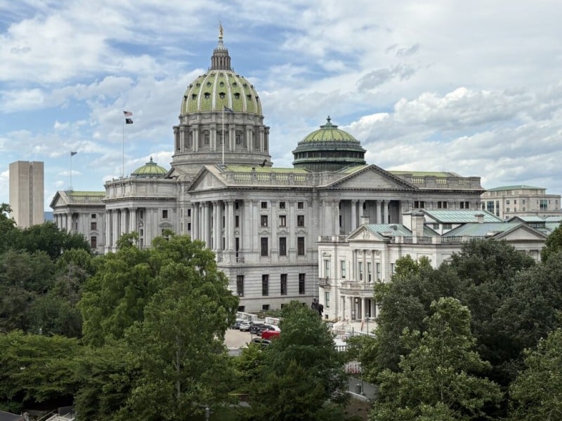 The Pennsylvania State Capitol building with its prominent green dome rises behind a line of dense green trees.