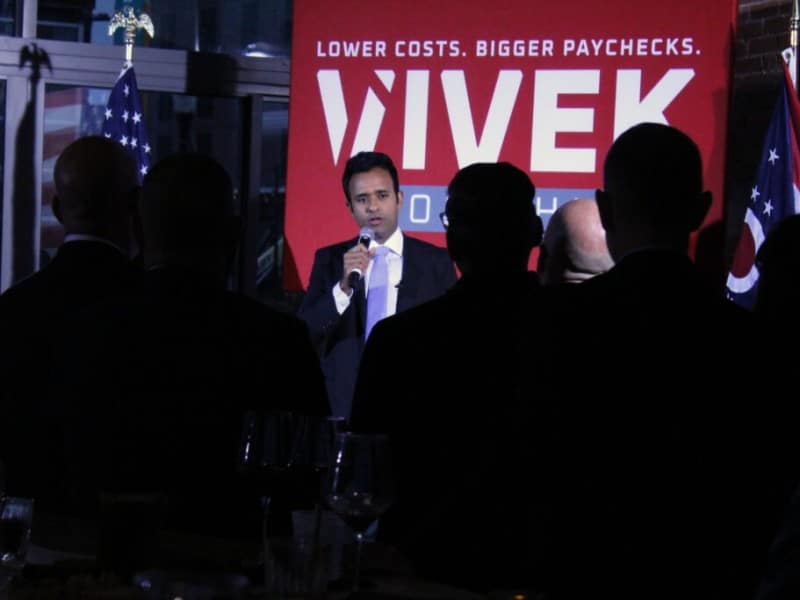 Vivek Ramaswamy speaks to an audience in front of a campaign sign reading "LOWER COSTS. BIGGER PAYCHECKS. VIVEK."