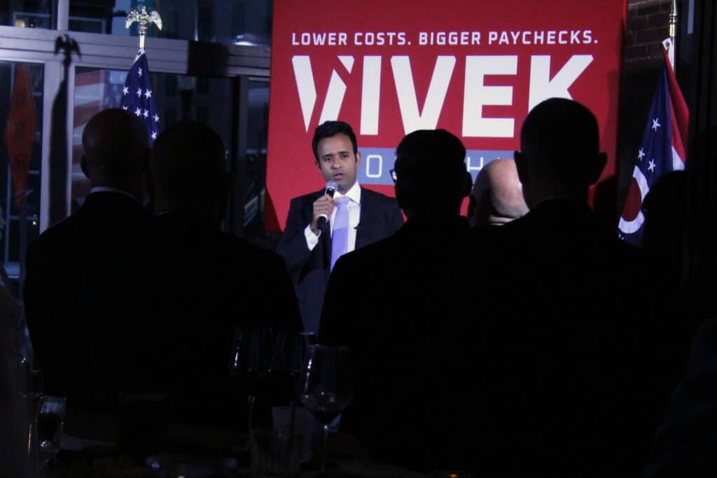 Vivek Ramaswamy speaks to an audience in front of a campaign sign reading "LOWER COSTS. BIGGER PAYCHECKS. VIVEK."