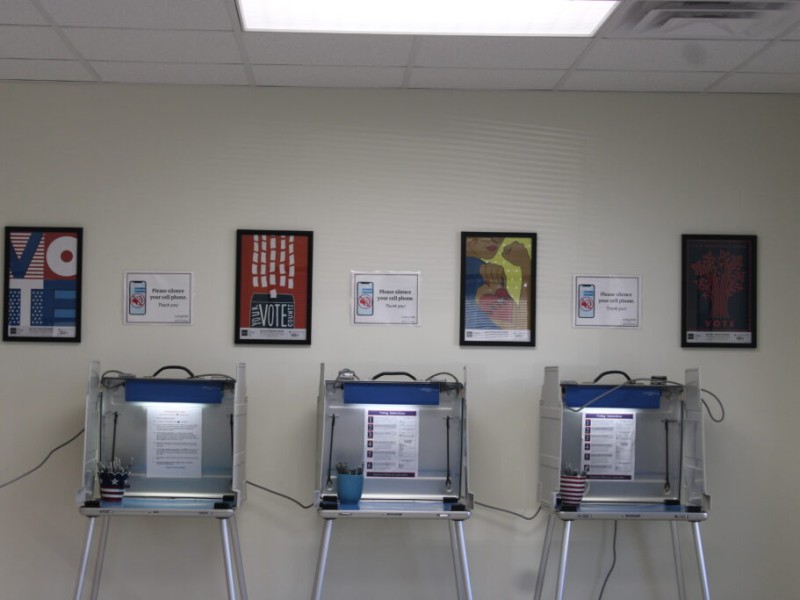 Three voting booths lined up against a wall beneath framed voting posters at a polling station.