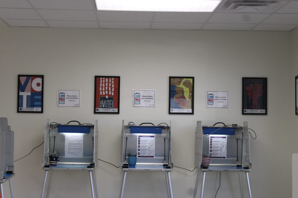 Three voting booths lined up against a wall beneath framed voting posters at a polling station.