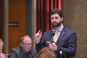 A man with a beard in a suit and tie gestures while speaking into a microphone in a legislative chamber.