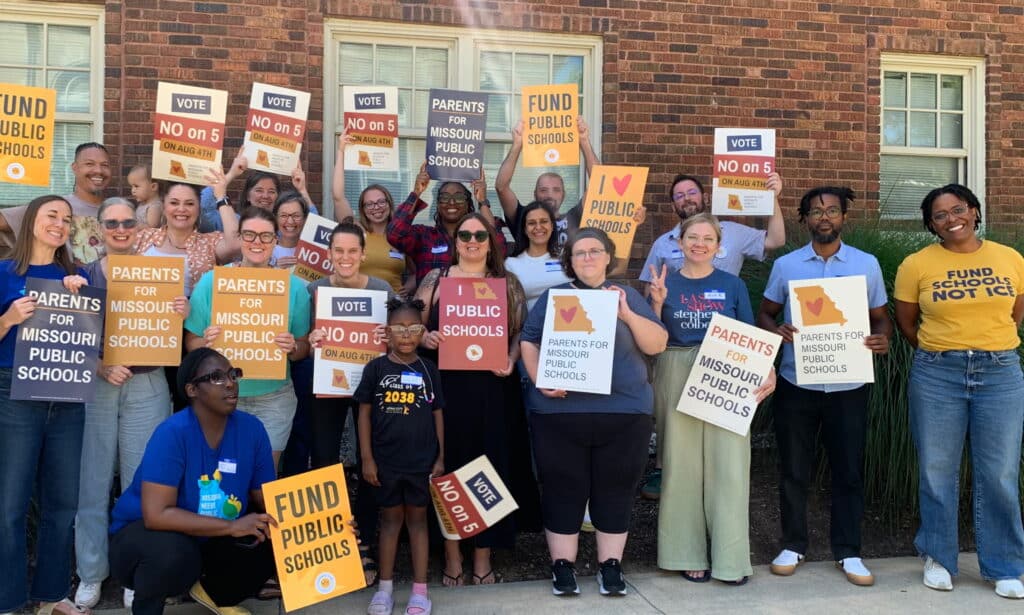 A group of people stand in front of a brick wall, holding signs that say "I love public schools," "Vote no on 5" and "Fund Public Schools."