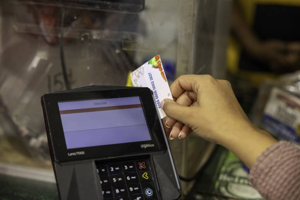 A hand slides a WIC Farmers' Market Benefit card, decorated with illustrations of vegetables, into the slot of a black Ingenico electronic payment terminal. The person making the transaction wears a long-sleeved, textured mauve sweater.