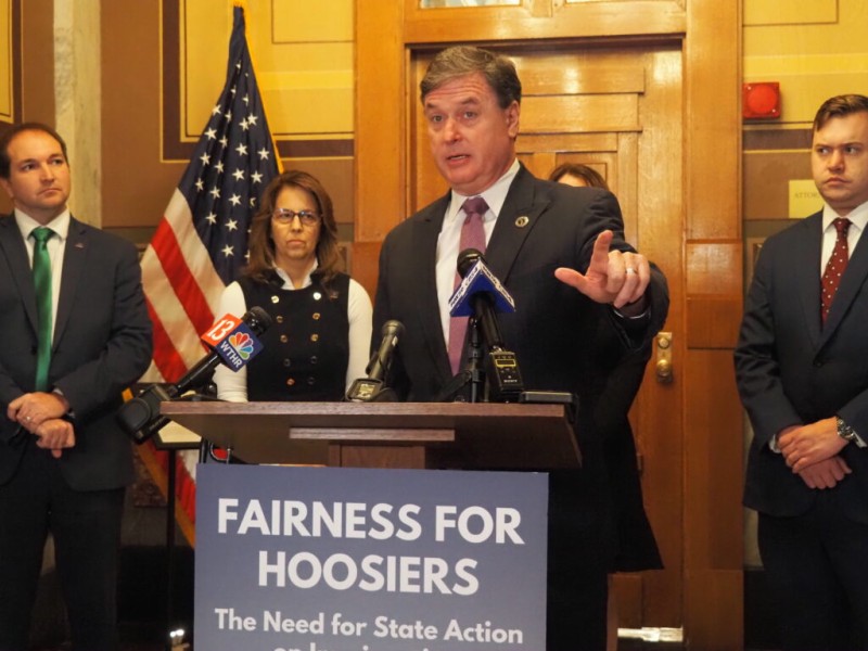A man stands at a wooden podium speaking into multiple microphones and gesturing with his hand during a press conference featuring a sign that reads "FAIRNESS FOR HOOSIERS." He wears a dark suit, white shirt, and a patterned purple tie, while the three colleagues standing behind him are also dressed in professional business suits and attire.