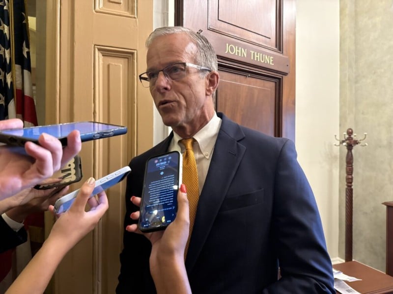U.S. Senate Majority Leader John Thune, R-S.D., stands outside an office door bearing his name, speaking to a huddle of reporters holding out smartphones to record his remarks. He wears a dark navy blue suit jacket over a crisp white dress shirt, paired with a vibrant gold textured tie and wire-rimmed glasses.