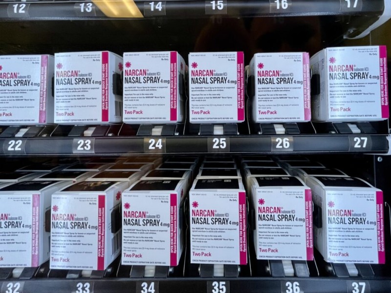 Multiple rows of white and pink boxes of Narcan (naloxone HCl) Nasal Spray 4 mg are lined up inside a vending machine dispensed for public access. Each two-pack box is positioned neatly over a numbered slot, ranging sequentially from 12 through 17 on the top shelf and 22 through 27 and 32 through 37 on the lower shelves.