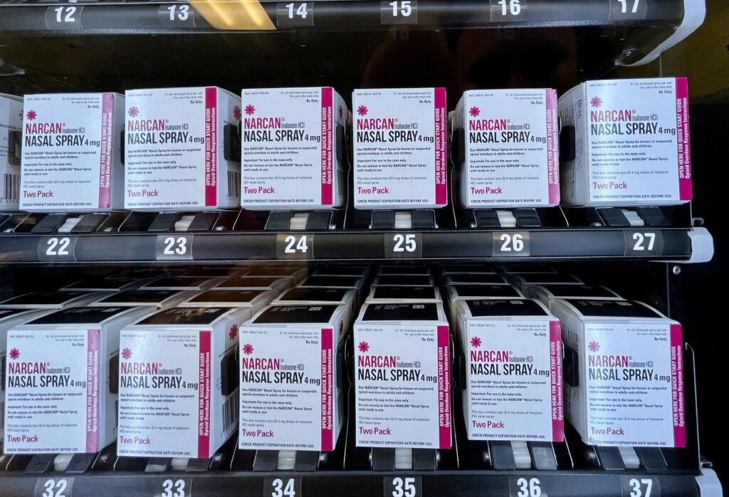 Multiple rows of white and pink boxes of Narcan (naloxone HCl) Nasal Spray 4 mg are lined up inside a vending machine dispensed for public access. Each two-pack box is positioned neatly over a numbered slot, ranging sequentially from 12 through 17 on the top shelf and 22 through 27 and 32 through 37 on the lower shelves.