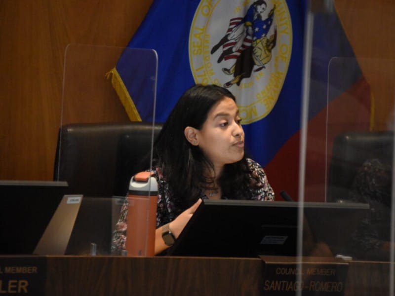 A woman, Gabriela Santiago-Romero, sits at a dais behind a clear plastic barrier, speaking while gesturing slightly during a public meeting. She is wearing a dark floral patterned top and a smartwatch, with a nameplate in front of her reading "COUNCIL MEMBER SANTIAGO-ROMERO" and the Wayne County, Michigan flag draped behind her.