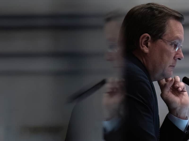 Tennessee House Speaker Cameron Sexton is captured in a sharp profile view, looking contemplative with his hand resting against his chin as a blurred, ghosted reflection of his face appears to the left. He is wearing a dark suit jacket over a white collared shirt, along with wire-rimmed glasses, while seated in front of a microphone.