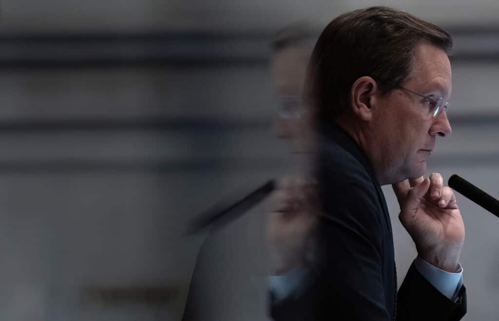 Tennessee House Speaker Cameron Sexton is captured in a sharp profile view, looking contemplative with his hand resting against his chin as a blurred, ghosted reflection of his face appears to the left. He is wearing a dark suit jacket over a white collared shirt, along with wire-rimmed glasses, while seated in front of a microphone.