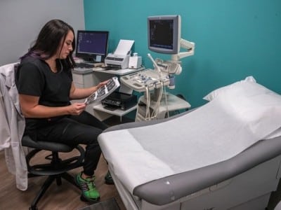 A medical worker sits on a rolling stool in an examination room, carefully reviewing a sheet of ultrasound images. She is wearing black scrubs and bright green running shoes, with a white lab coat draped over the back of her chair next to an ultrasound machine and an examination table.