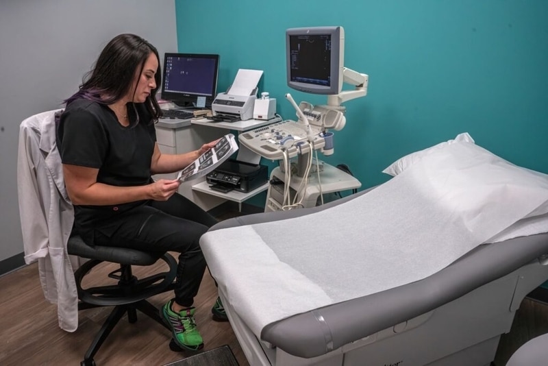 A medical worker sits on a rolling stool in an examination room, carefully reviewing a sheet of ultrasound images. She is wearing black scrubs and bright green running shoes, with a white lab coat draped over the back of her chair next to an ultrasound machine and an examination table.