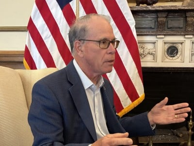 Indiana Governor Mike Braun sits in an armchair and gestures with his hands while speaking during an interview in an office setting. He is wearing a blue textured blazer over a light-colored button-down shirt, dark trousers and tortoiseshell-framed glasses, with an American flag positioned behind him.