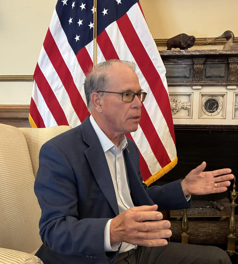 Indiana Governor Mike Braun sits in an armchair and gestures with his hands while speaking during an interview in an office setting. He is wearing a blue textured blazer over a light-colored button-down shirt, dark trousers and tortoiseshell-framed glasses, with an American flag positioned behind him.