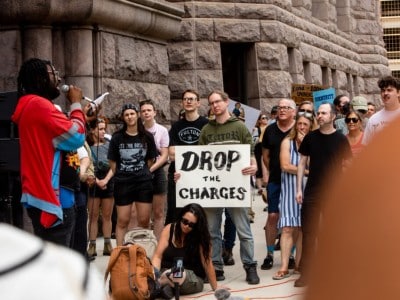 A speaker with long dreadlocks addresses a crowd outside a large stone building during a protest in Minneapolis. A demonstrator stands at the center holding a white sign that reads "DROP THE CHARGES" in bold black lettering, wearing a green hooded sweatshirt and jeans.