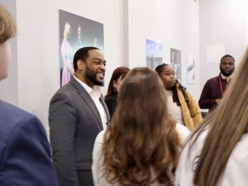 A smiling Charles Booker stands inside an event space or gallery, engaging in conversation with a group of people in front of framed photographs hanging on the wall. He is wearing a gray suit jacket over an open-collared white shirt, while the others in the crowd are dressed in a mix of professional and casual attire.