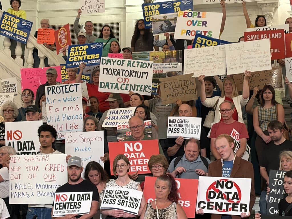 A large crowd of protestors fills the steps inside a building, holding numerous signs opposing data centers and AI development, with prominent messages including "DAY OF ACTION MORATORIUM ON DATA CENTERS" and "PEOPLE OVER PROFITS." The diverse group of activists wears various casual clothing, such as T-shirts, tank tops and button-downs.
