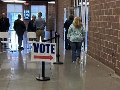 A sign reading "VOTE" with a red arrow stands in the foreground of a spacious hallway as voters arrive at a polling location. A woman in the foreground walks toward the entrance wearing a blue and white patterned blouse with blue jeans, while others nearby are dressed in casual shirts, trousers and jackets.