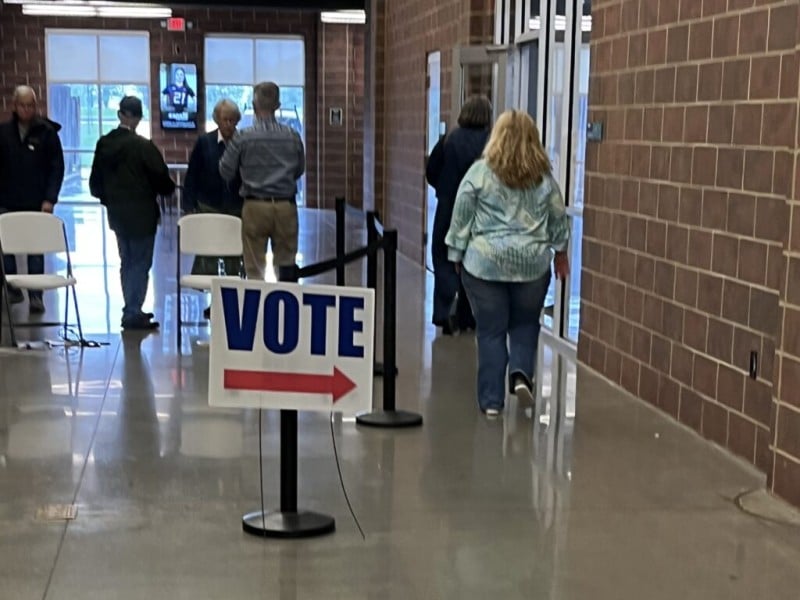 A sign reading "VOTE" with a red arrow stands in the foreground of a spacious hallway as voters arrive at a polling location. A woman in the foreground walks toward the entrance wearing a blue and white patterned blouse with blue jeans, while others nearby are dressed in casual shirts, trousers and jackets.