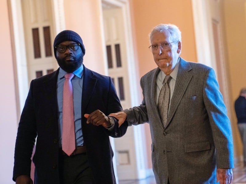 U.S. Senator Mitch McConnell walking in the U.S. Capitol with assistance from an aide.