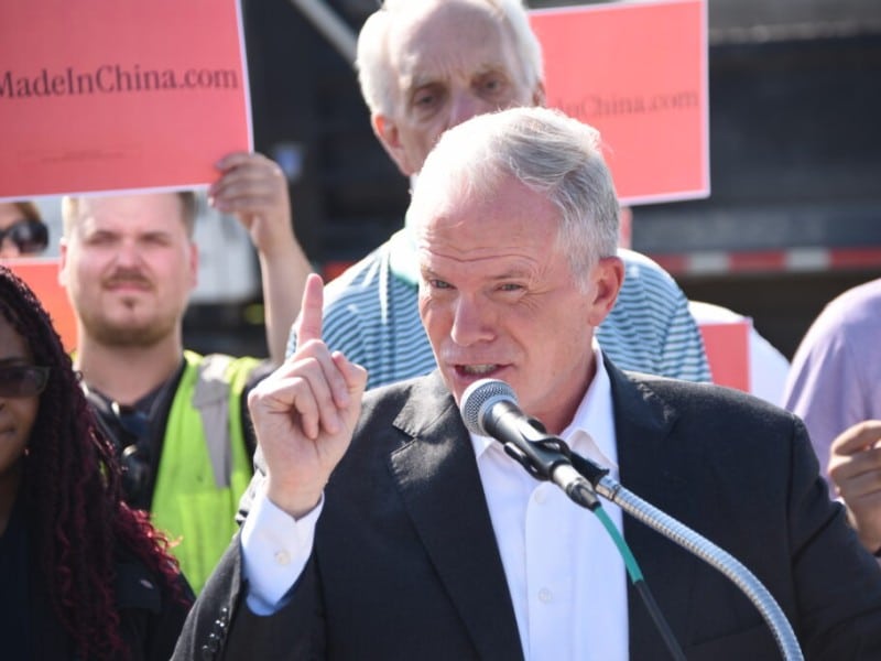 Mike Cox speaks into a microphone while raising his index finger during an outdoor press conference or rally, surrounded by supporters holding red signs. He is wearing a dark suit jacket over a white button-down collared shirt.