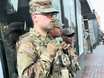 A line of three Michigan National Guard members stands on a brick sidewalk alongside a building's glass storefront. They are wearing matching camouflage utility uniforms and patrol caps, complete with tactical vests, tan combat gloves, and communication gear.